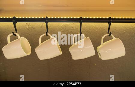White coffee cups hanging on hooks of the grey kitchen wall. Live kitchen. Stock Photo
