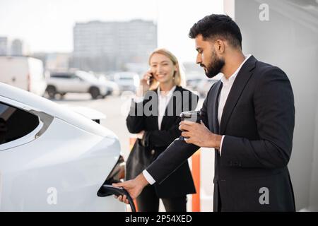 Side view of bearded indian businessman plugging charging cord into electric car while caucasian woman having mobile conversation. Two office workers using modern transport with zero emission. Stock Photo