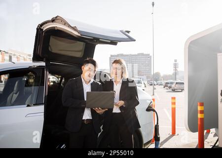 Two focused multiracial business partners in formal attire working on wireless laptop while waiting for luxury electric car to be charged. Concept of people, ecology and technology. Stock Photo