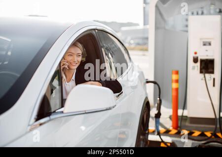 Successful caucasian business lady in stylish black suit sitting inside charging electric car and using modern smartphone for conversation. Technology and transport concept. Stock Photo