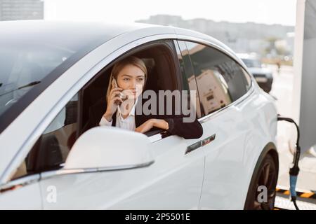 Successful caucasian business lady in stylish black suit sitting inside charging electric car and using modern smartphone for conversation. Technology and transport concept. Stock Photo