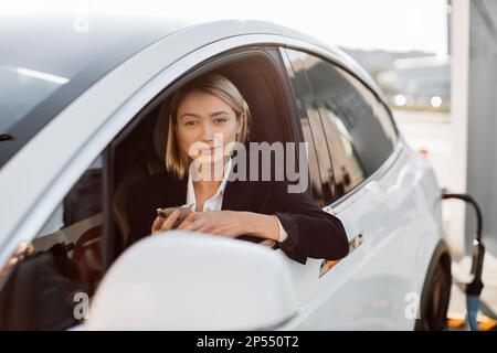 Attractive caucasian woman with modern smartphone in hands sitting inside luxury car while recharging battery on auto station. Business lady in stylish suit working online during trip break. Stock Photo
