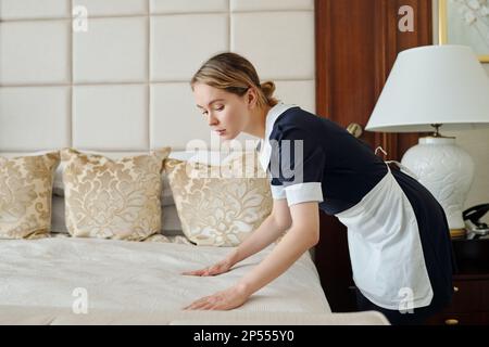 Side view of young housemaid putting clean bedclothes on double bed while preparing room of luxurious five star hotel for new travelers Stock Photo