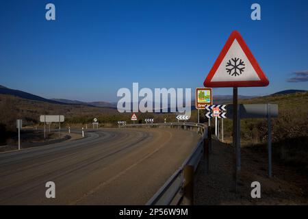 Dangerous curve with tire tracks with warning sign for snow in sunny winter day Stock Photo