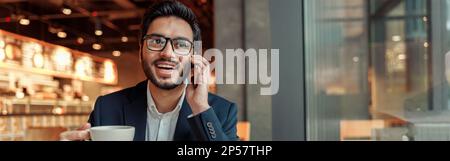 Handsome indian businessman in glasses is talking phone with client during lunch time in cozy cafe Stock Photo