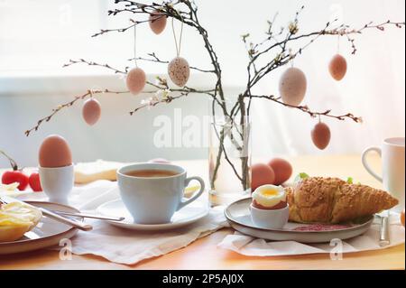 Easter breakfast in the morning sun with coffee and croissant on a table at the window, decoration from natural eggs hanging on spring branches, copy Stock Photo