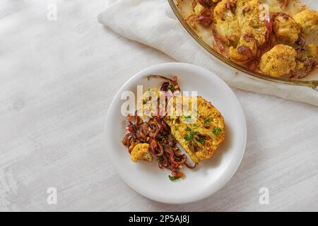 Baked spicy cauliflower steaks with red onions and parsley garnish on a white plate, healthy vegetable dish for low carb diet, high angle view from ab Stock Photo