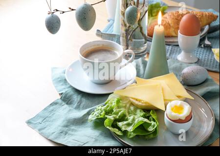 Breakfast table setting with bread roll, cheese, boiled egg and coffee on a pastel turquoise napkin, hanging Easter eggs and candle as decoration, cop Stock Photo