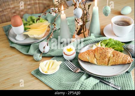 Breakfast with Easter eggs, croissant, coffee and sage green decoration with candles on a wooden table, copy space, selected focus, narrow depth of fi Stock Photo