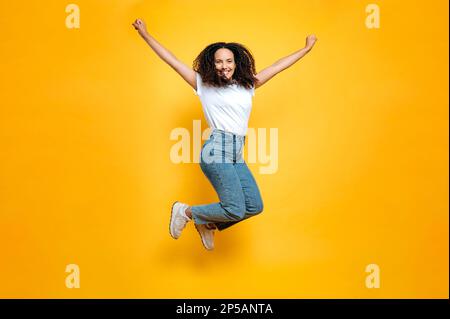 Full length photo of cheerful mixed race young woman with curly hair dressed in casual clothes, bobbing high on isolated orange background, looking at camera, smiling happily Stock Photo