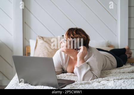 Smiling middle aged plus size woman lying on bed, working on laptop from home Stock Photo