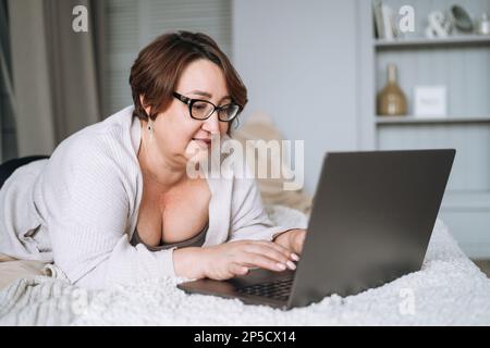 Smiling middle aged plus size woman lying on bed, working on laptop from home Stock Photo
