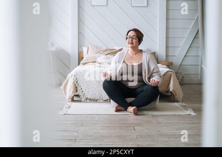 Middle aged plus size smiling brunette woman in home wear practicing yoga in bedroom Stock Photo