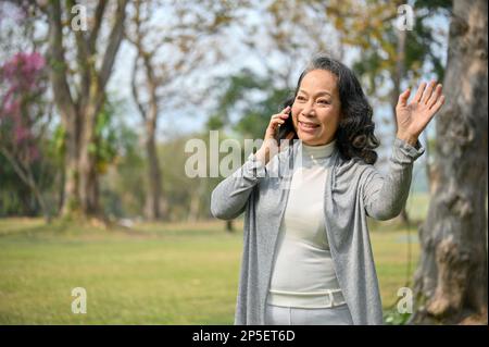 Happy aged Asian woman in casual clothes waving her hand and making a hello gesture, bumped into her neighbor in the green park while talking on the p Stock Photo