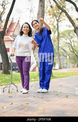 Portrait of a caring Asian female caregiver or nurse assistant enjoys talking and walking in the green park with an old lady patient. Stock Photo