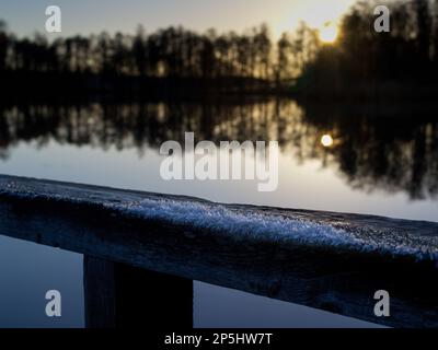 The sun goes down behind the trees and is reflected in the lake with the frost on the railing in for Stock Photo