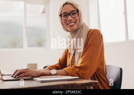 Woman wearing hijab working from home. Woman working on laptop looking at camera and smiling. Stock Photo