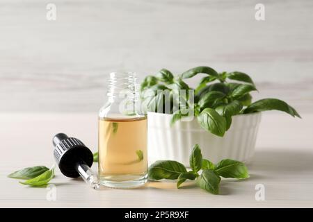 Glass bottle of basil essential oil near bowl with leaves on white wooden table Stock Photo