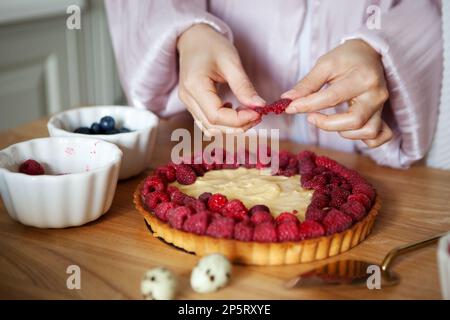 Close up of woman's hands decorating raspberry tart, cooks delicious pie with fresh berries and cream cheese Stock Photo