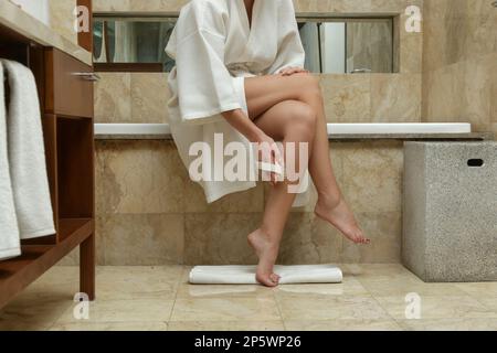Portrait of young woman in white cotton bathrobe dry brushing legs using massage brush in bathroom Stock Photo