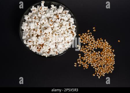 Ready-to-eat popcorn in a glass bowl and corn kernels on a black background, top view and copy space. Stock Photo