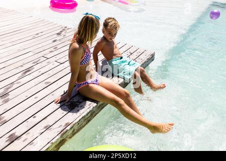 Happy biracial brother and sister sitting by the swimming pool Stock Photo