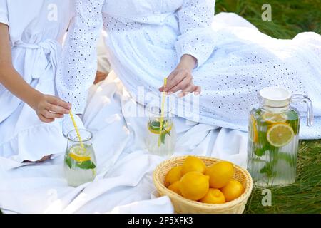 close up hands of mother and daughter drink lemonade picnic on green grass in summer Stock Photo