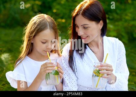 girl child holds glass of lemonade in hands and drinks on picnic with mother outdoors Stock Photo