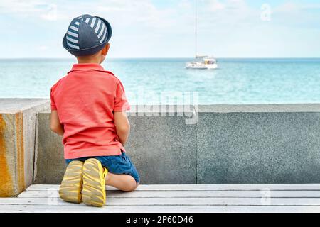 Dreaming little boy with panama looks at yacht sailing on sea from bench on seafront backside view. Toddler kid rests on city embankment Stock Photo