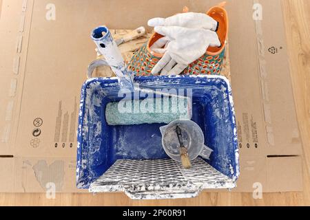 Blue painting square plastic bucket standing on cardboard, dirty from white dry paint including basic tools like grid, roller, measuring jug, brush Stock Photo