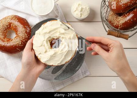 Woman making bagel with cream cheese at white wooden table, top view Stock Photo