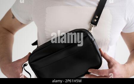 Side view man in a white t-shirt with a leather black handmade bag over his shoulder. Dark designer banana bag. Comfortable small bag for walking. Stock Photo