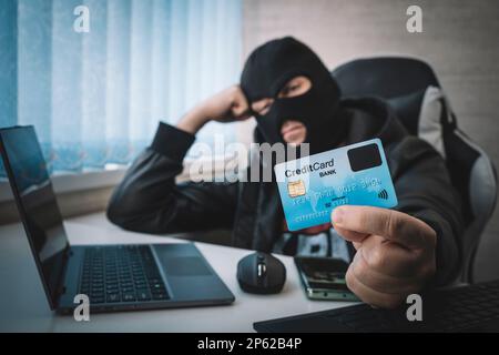 Internet Theft - a man wearing a balaclava and holding a credit card while sat behind a laptop, white background Stock Photo