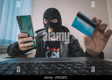 Male hacker in a robber mask uses phone, credit card and laptop in some fraudulent scheme. Cyber thief stole the personal data and credit card informa Stock Photo