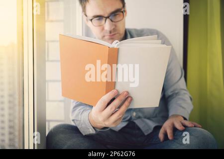 Young man sitting next to a window in cafe, reading a book and enjoying his leisure time Stock Photo