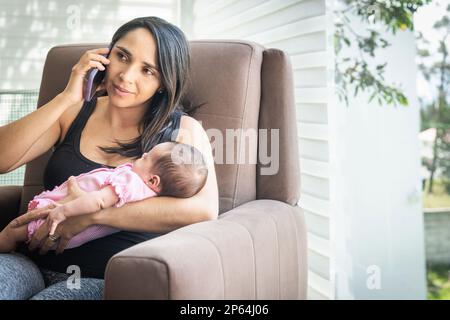 Latin woman sitting on a couch working on her cell phone with her sleeping baby at home Stock Photo