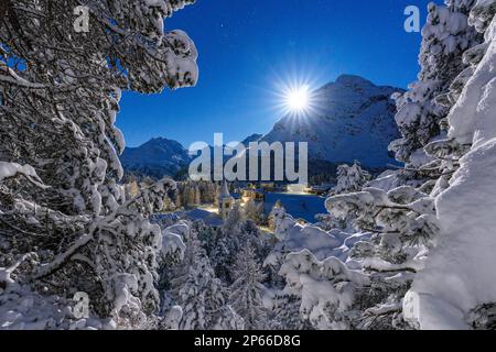 Moon glowing over the snowy bell tower of Chiesa Bianca and woods in winter, Maloja, Bregaglia, Engadine, Canton of Graubunden, Switzerland, Europe Stock Photo