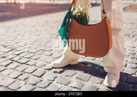 close up of female leather handbag with tied scarf and shoes woman holds brown purse with bouquet of white spring flowers outdoors stylish accessori 2p67jb5
