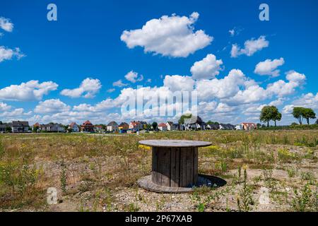 An empty cable spool lying on a wasteland. Houses in the background under a blue sky with light clouds. Stock Photo