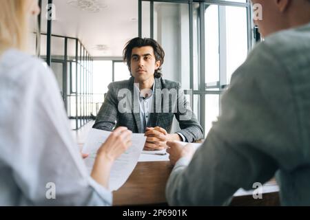Portrait of confident male job seeker in formal wear interviewed by unrecognizable two HR managers advisors at meeting in corporate office. Stock Photo