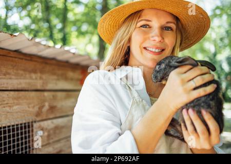Close up of a young beautiful woman holding a small black rabbit. Caucasian pretty girl in a straw hat and a white shirt smiles and holds a cute bunny Stock Photo