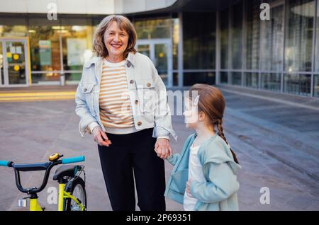Young grandmother teaches her cute little granddaughter to ride a bicycle. Caucasian woman in glasses and casual clothes smiles at a little girl and s Stock Photo