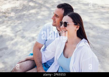 Caucasian couple in love on a sandy beach. Young man lovingly looks at a beautiful woman in sunglasses. Girlfriend and boyfriend smile at each other. Stock Photo