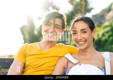 Handsome man wearing yellow tshirt and blue swimsuit sits with beautiful woman in dungarees in a park in a sunny day during holidays. Young adults cou Stock Photo