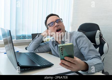 Tired bored businessman yawning at workplace near laptop, looking at smartphone, checking time, boring work, dead end job, overwork extra after hours, Stock Photo