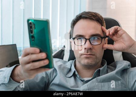 male use modern cellphone gadget, texting or messaging on wireless internet. Caucasian man in glasses sit at desk at home office look at smartphone sc Stock Photo