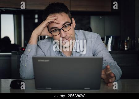 Young male concentrated guy adjusting eyeglasses while working on laptop and looking at pc screen during remote work from home office, overworking man Stock Photo