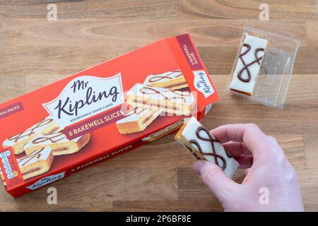 A man's hand holding a Mr Kipling Bakewell Slice with a packet of slices in the background. UK. Concept: snack food, unhealthy eating Stock Photo