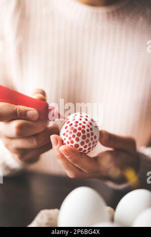 Unrecognizable woman painting  red dotted, colorful easter eggs, table top shot. Hands in focus. Stock Photo