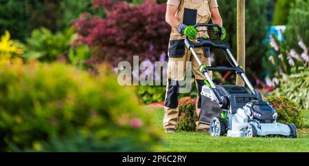 Summer Time Lawn Mowing in Colourful Blooming Garden. Professional Garden Worker Trimming the Grass. Stock Photo